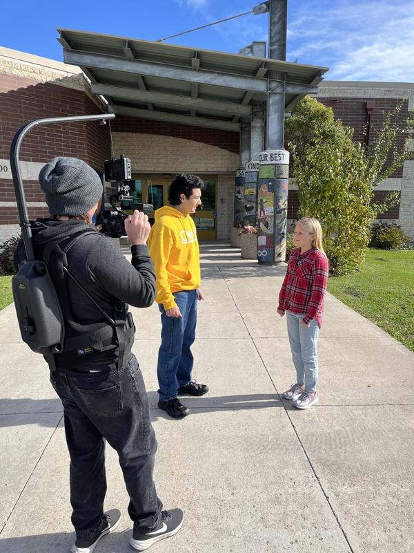 A student welcoming the host just outside of our school entrance, while being filmed by the camera.