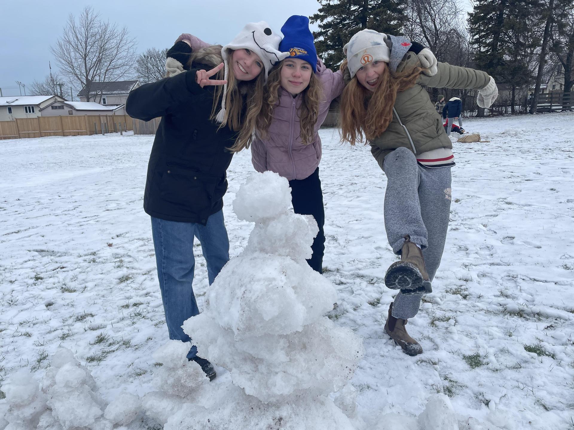 3 female students near snowman smiling and holding each other's shoulders