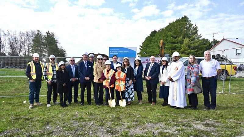 A large group of people standing together, smiling in hard hats and two students in the front holding shovels.