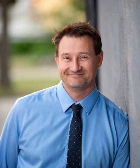 Gord Wood smiling and leaning against a wall wearing a blue dress shirt and black tie