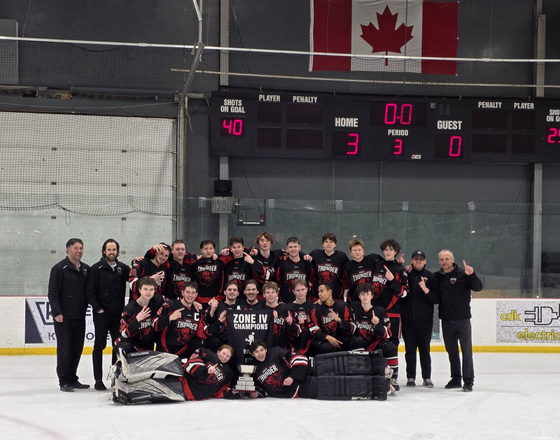 A hockey team celebrating with coaches on ice, holding trophies in front of a Canadian flag.
